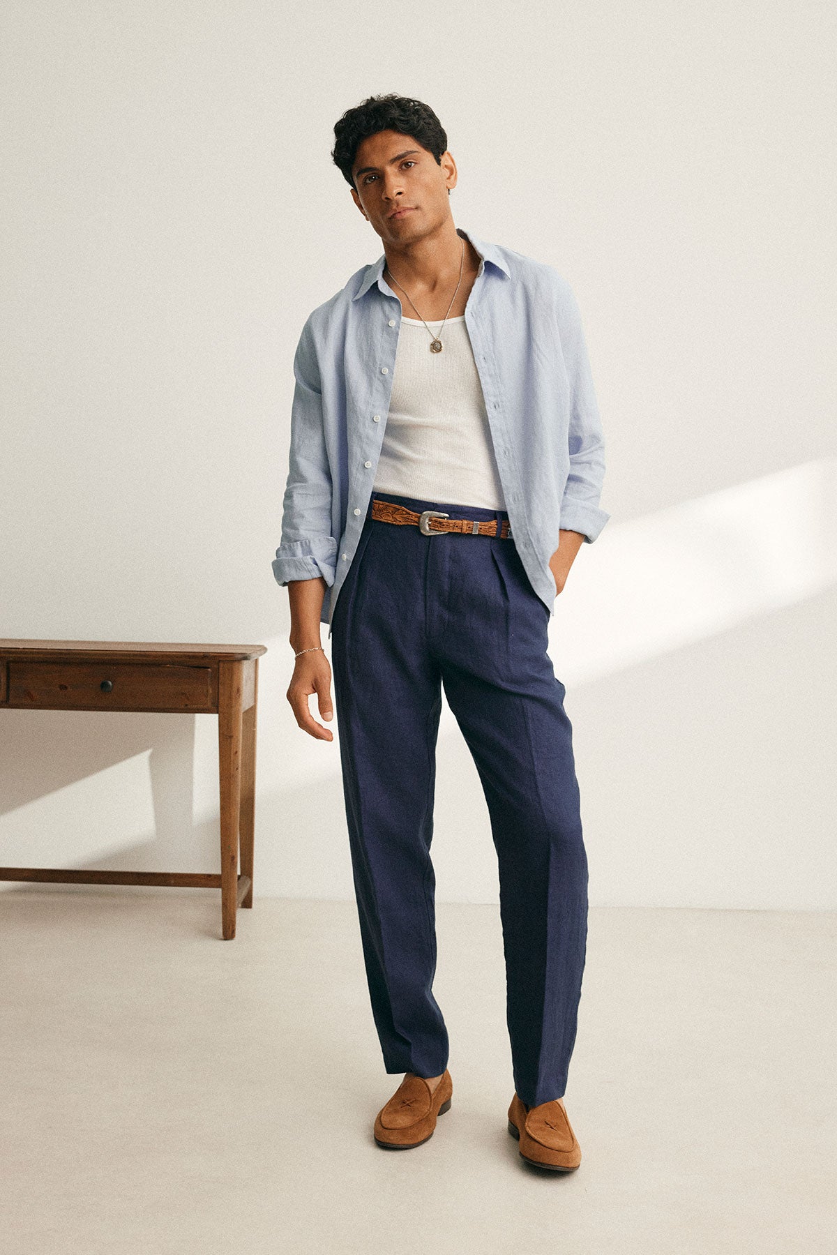   A man in a CARUSO LINEN BUTTON-UP SHIRT by Velvet by Graham & Spencer, layered over a white tank top and dark trousers, stands in a minimal room beside a wooden table, showcasing the versatility of his attire. 