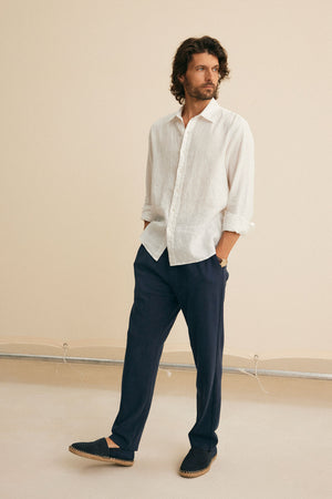 A man wearing the CARUSO LINEN BUTTON-UP SHIRT from Velvet by Graham & Spencer, paired with navy pants and espadrilles, stands against a neutral background, gazing to the side.