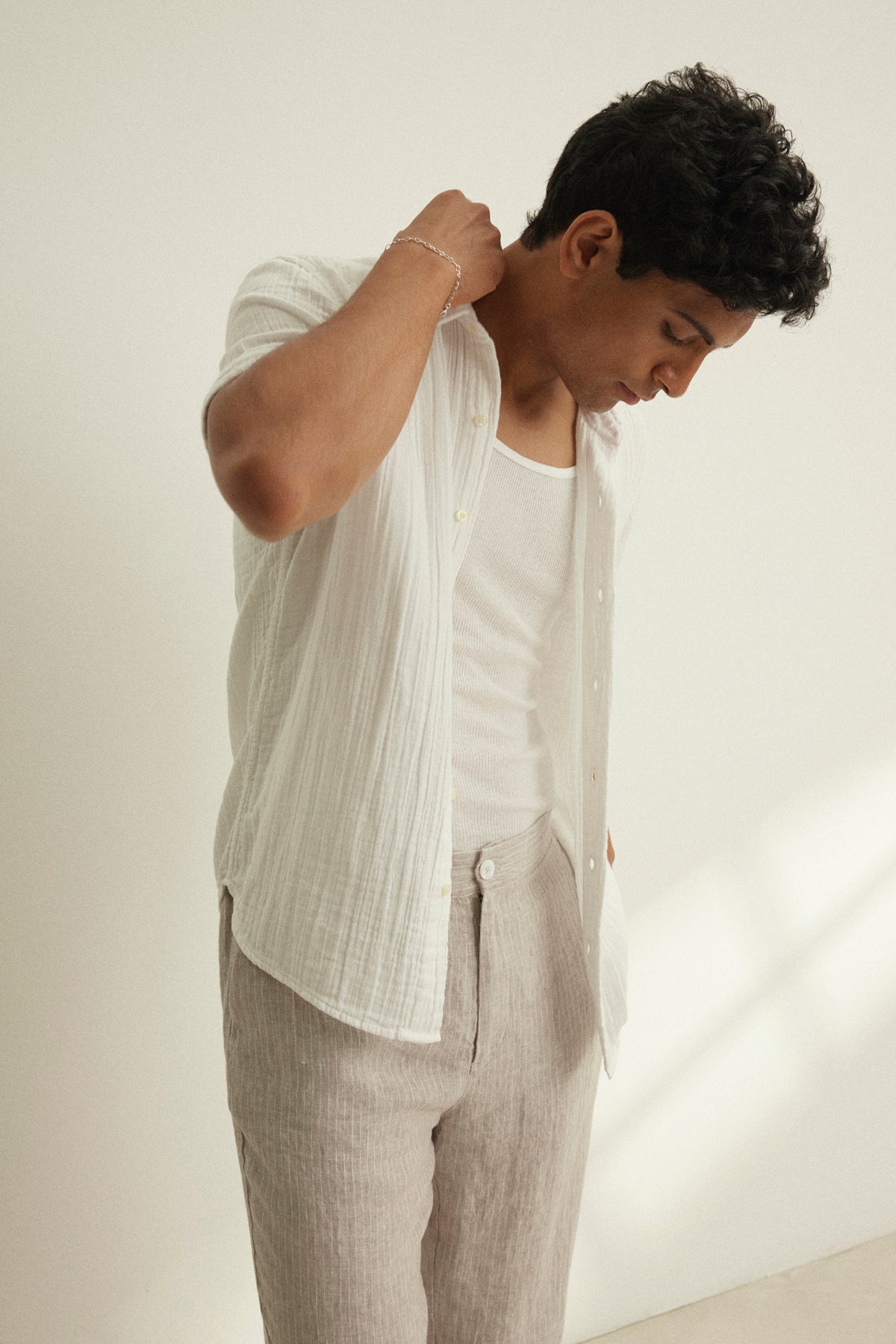 A man wears the CHRISTIAN COTTON GAUZE BUTTON-UP SHIRT by Velvet by Graham & Spencer over a white tank top with light beige pants, standing indoors and looking down with one hand behind his neck.-39152366158017