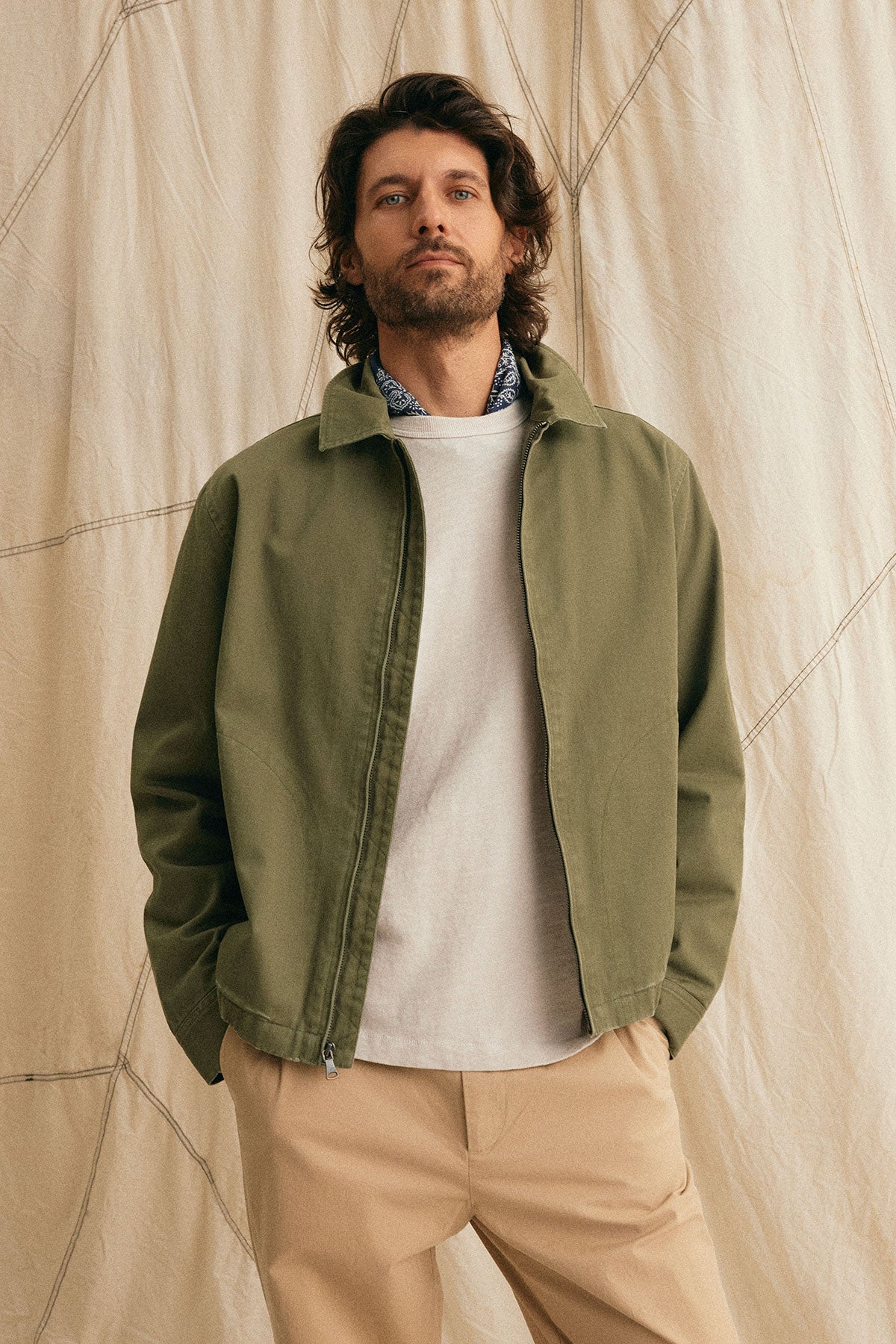   A man with wavy hair wears Velvet by Graham & Spencer's COLLINS JACKET, which has a plaid lining, over a white shirt and beige pants, standing against a cream draped backdrop. 