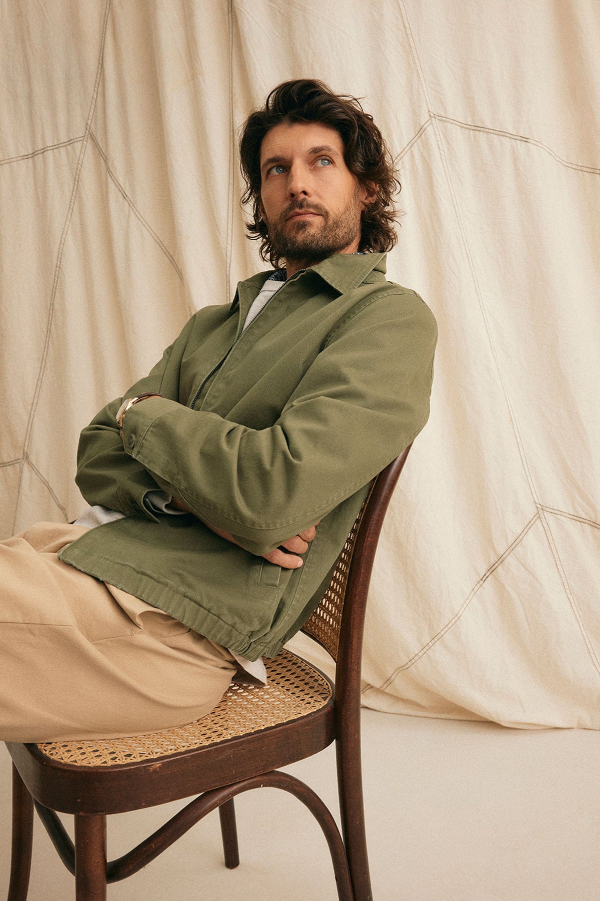   A bearded man sits on a chair in an olive green COLLINS JACKET from Velvet by Graham & Spencer, paired with beige pants. He gazes slightly left against a neutral backdrop with draped fabric, enhancing his timeless style. 