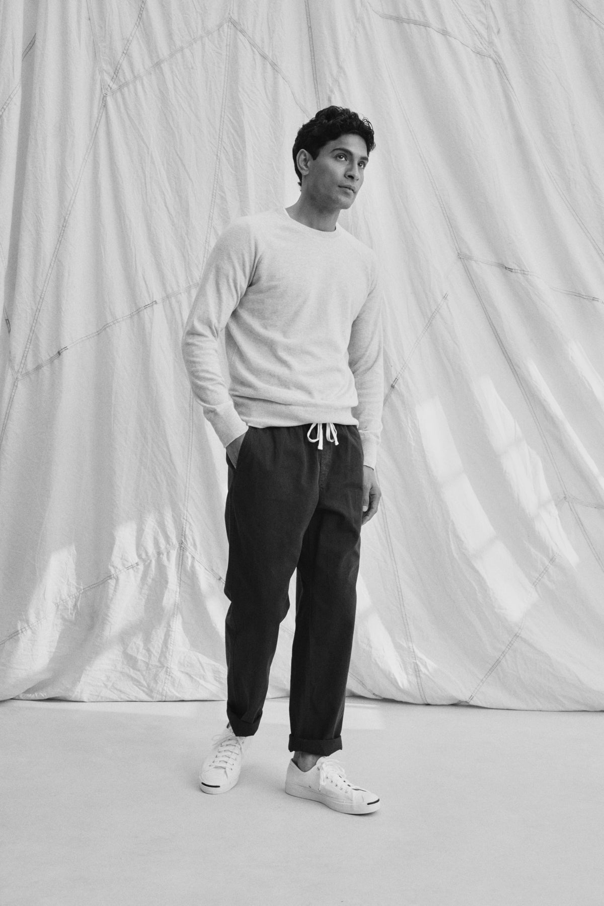 A person stands with hands in pockets, wearing a light sweater and BRANSON PANT by Velvet by Graham & Spencer. These cotton sanded twill drawstring pants feature an elastic waist and straight leg design. They sport sneakers against a draped fabric backdrop.-38643231948993