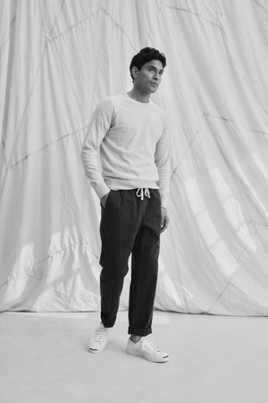 A person stands against a backdrop wearing the light-colored DARIO COTTON CASHMERE SWEATER by Velvet by Graham & Spencer, dark pants, and white sneakers, looking to the side.