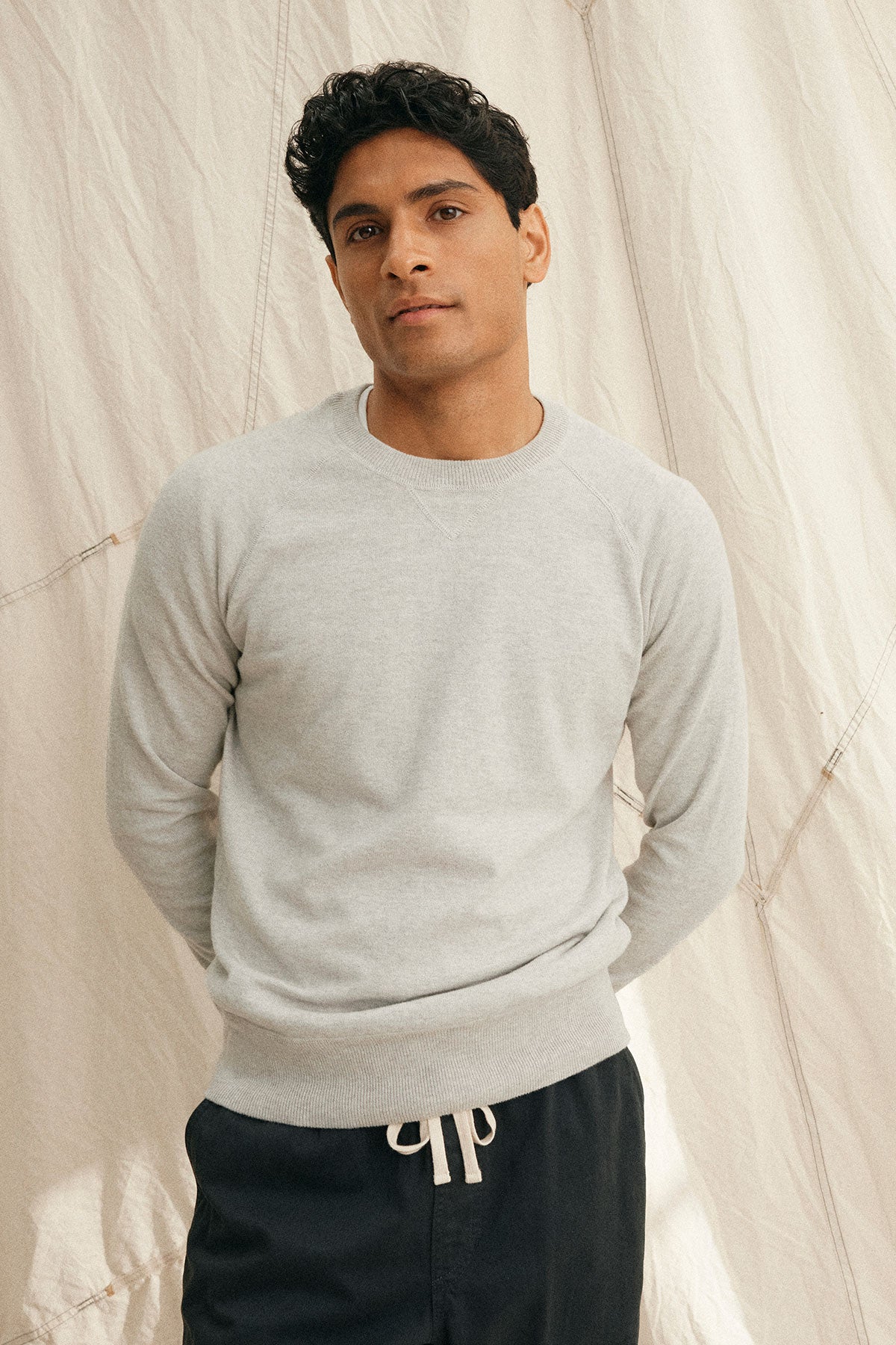 A person wearing a DARIO COTTON CASHMERE SWEATER by Velvet by Graham & Spencer in light gray and dark pants stands with hands behind their back against a beige fabric backdrop.-38877188325569