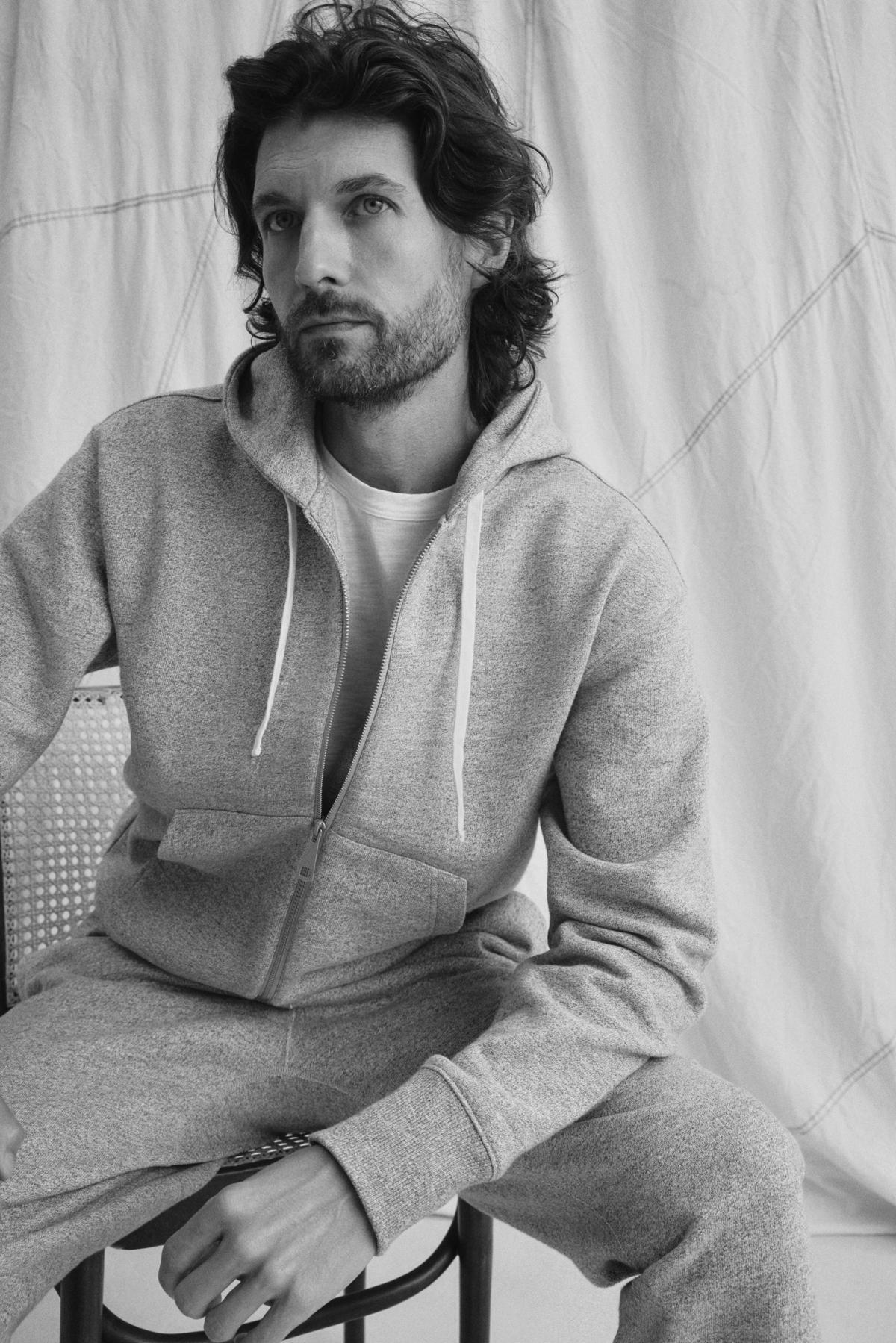   A person sits in a black-and-white image wearing the DRACO HOODIE by Velvet by Graham & Spencer, paired with pants, as they gaze to the side against a backdrop of draped fabric. 