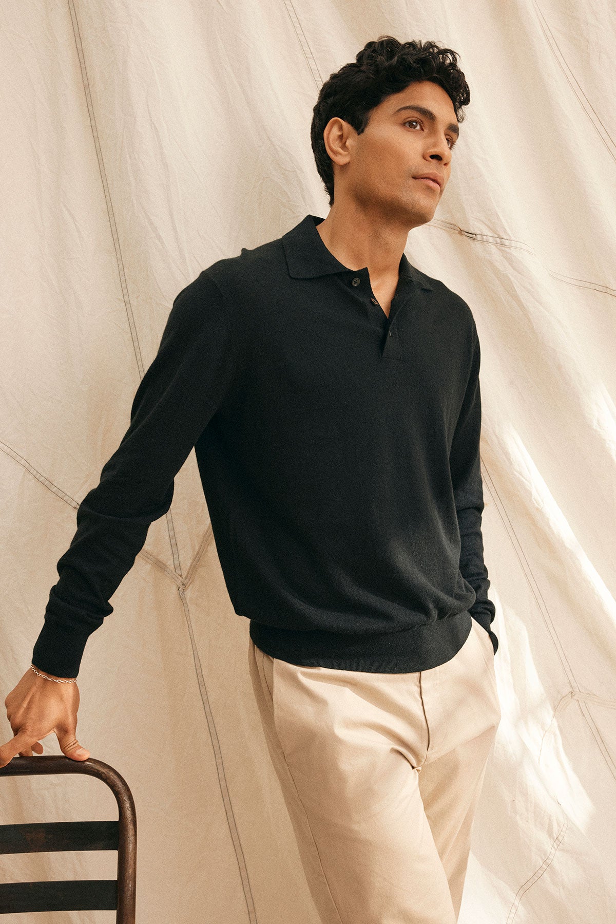 Wearing the MATTEO COTTON LINEN SWEATER POLO by Velvet by Graham & Spencer, this individual in a black long-sleeve shirt and beige pants leans against a chair, gazing upward. A draped white fabric backdrop accentuates the outfit's lightweight comfort and style.-38877192192193