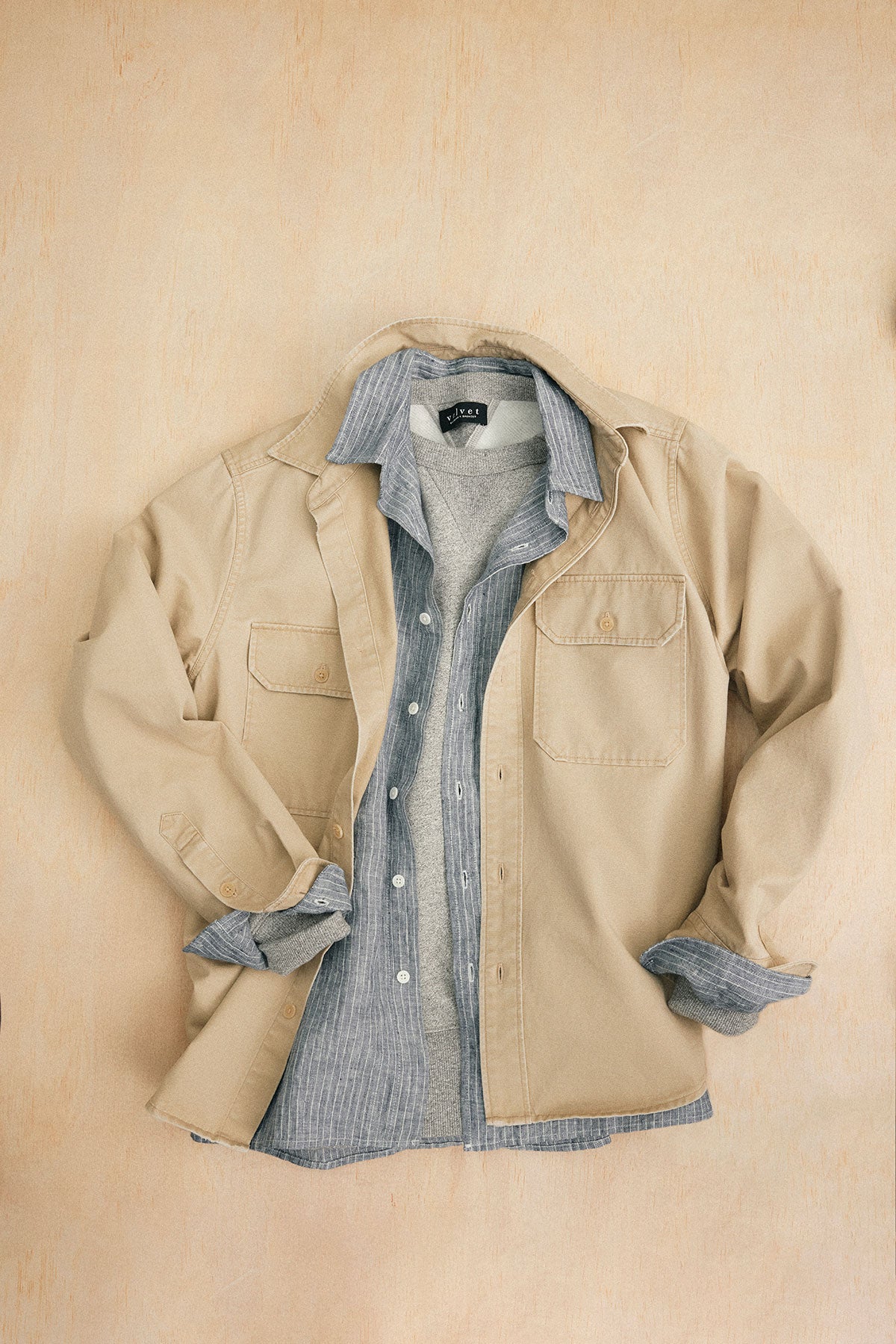 The ORLANDO BUTTON-UP SHIRT from Velvet by Graham & Spencer, in beige with chest flap pockets, is stylishly displayed layered over a gray striped shirt and light gray sweater on a flat surface for a structured fit.-38877236068545