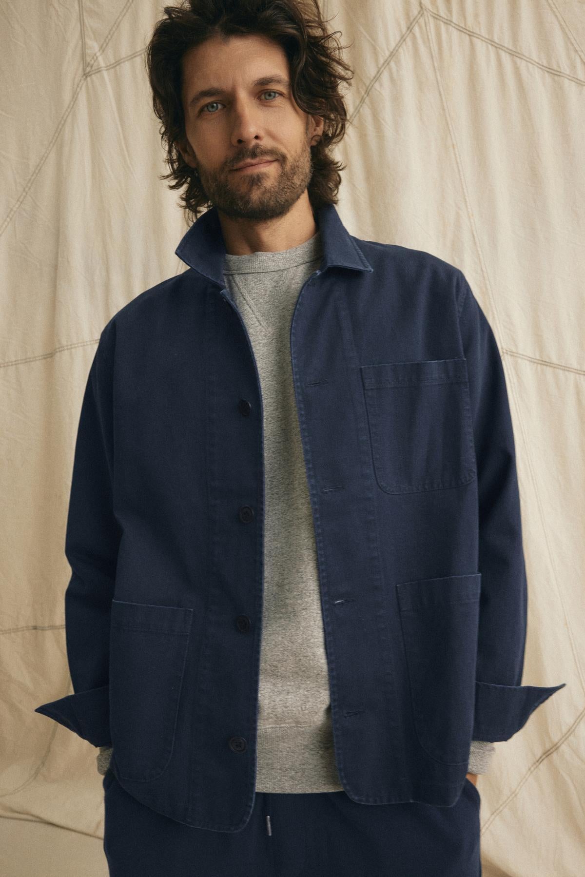   A man with shoulder-length hair models the BLAYNE JOGGER by Velvet by Graham & Spencer, wearing a navy jacket over a gray shirt in front of a cream-colored fabric backdrop. 