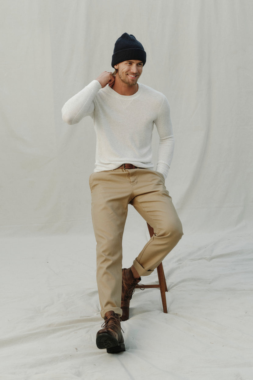 A man sitting on a stool wearing a Velvet by Graham & Spencer AUGUSTUS RIB KNIT CREW beanie and khaki pants.