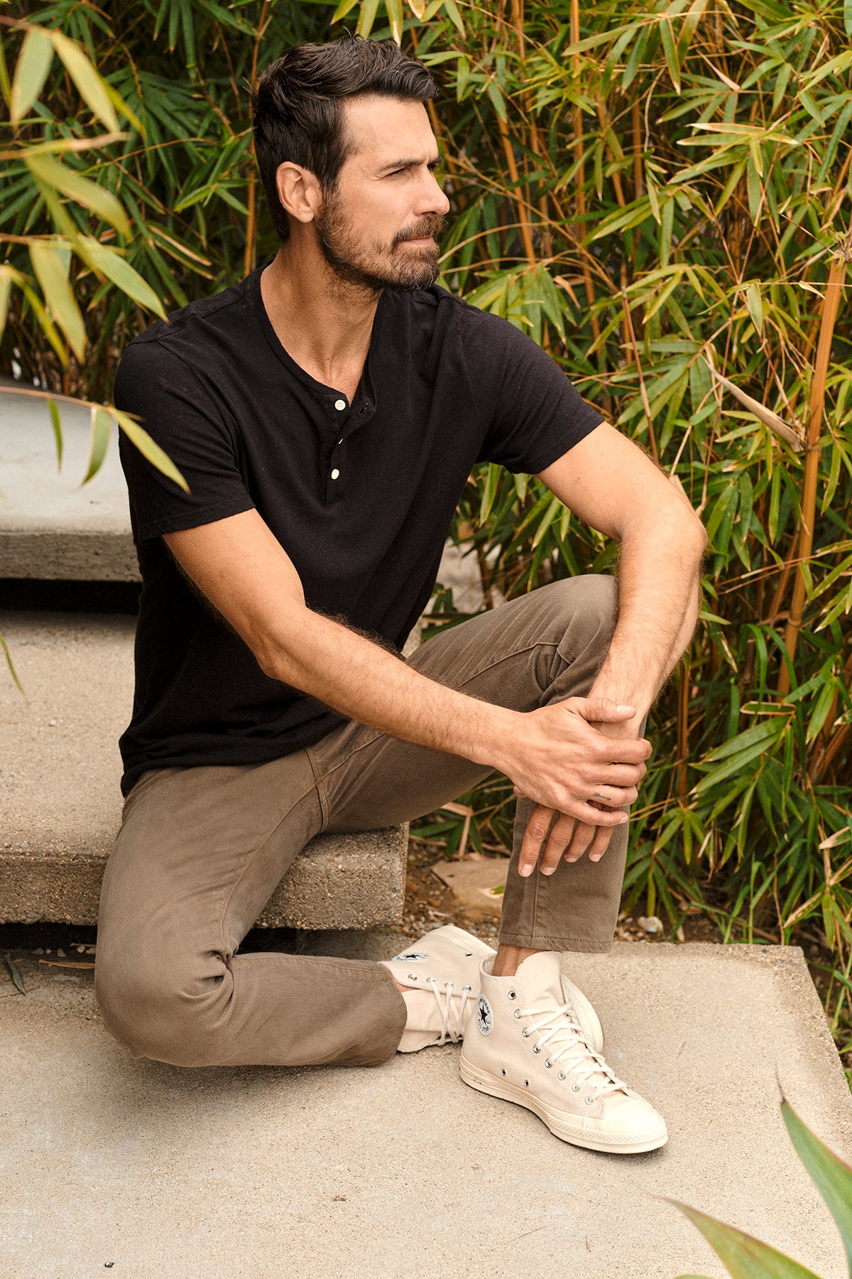 Man wearing Lionel Henley in black sitting outdoors on cement steps with greenery, brown pants and white Converse-26468356456641