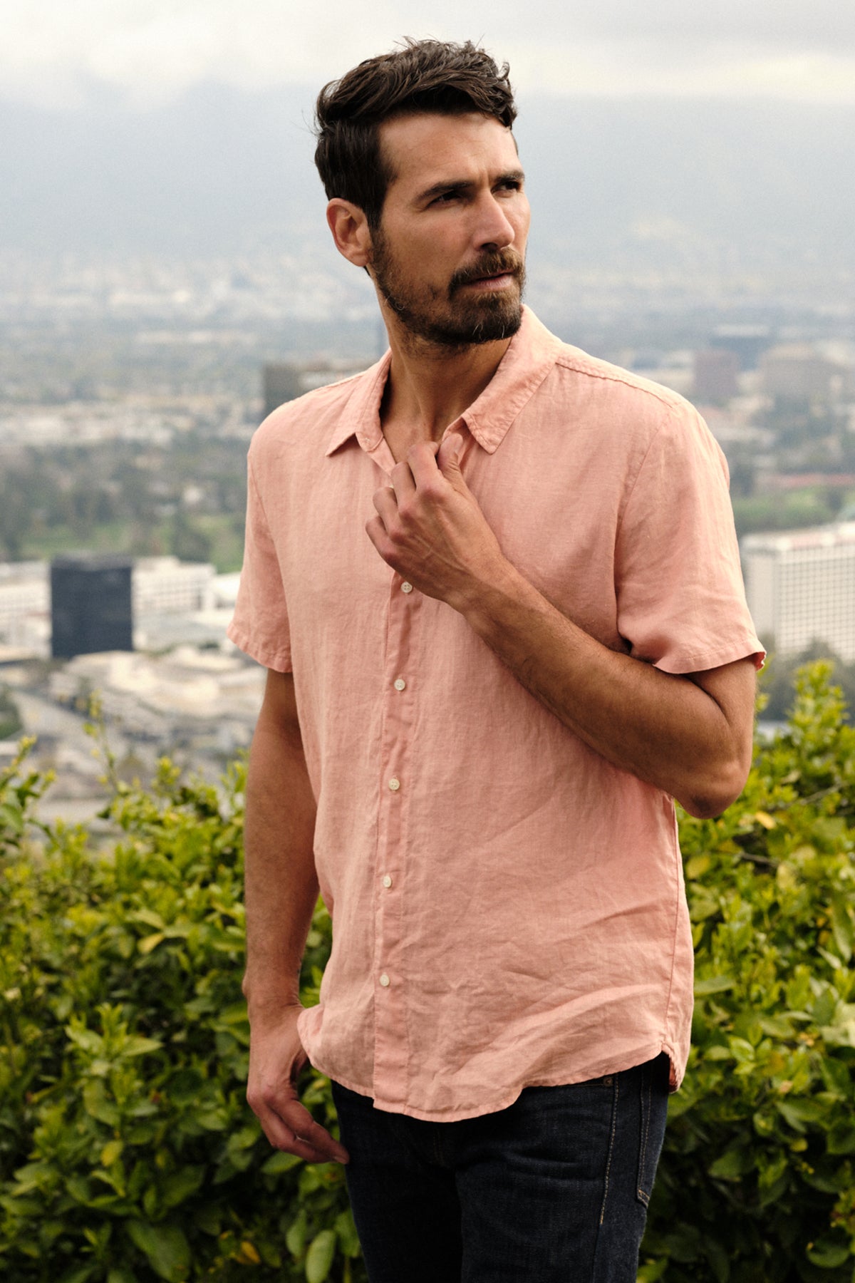   Mackie Button-Up Shirt in bronze with dark denim, model standing outdoors near greenery overlooking downtown 