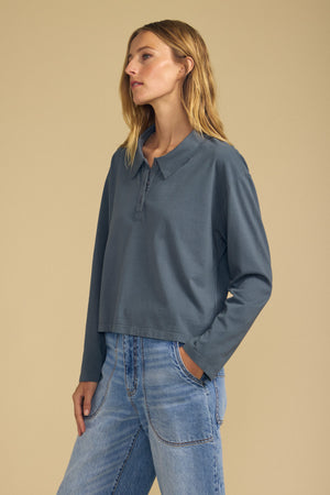 A woman stands against a beige background, wearing the Velvet by Graham & Spencer NILLI POLO—a loose-fitting, boxy silhouette blue long-sleeve collared shirt—paired with light blue jeans, her hand casually in her pocket.