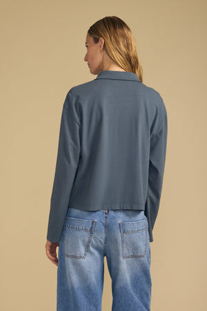 A person stands facing away, wearing the NILLI POLO by Velvet by Graham & Spencer—a gray, boxy-fit polo with drop shoulders—paired with light blue jeans against a beige background.