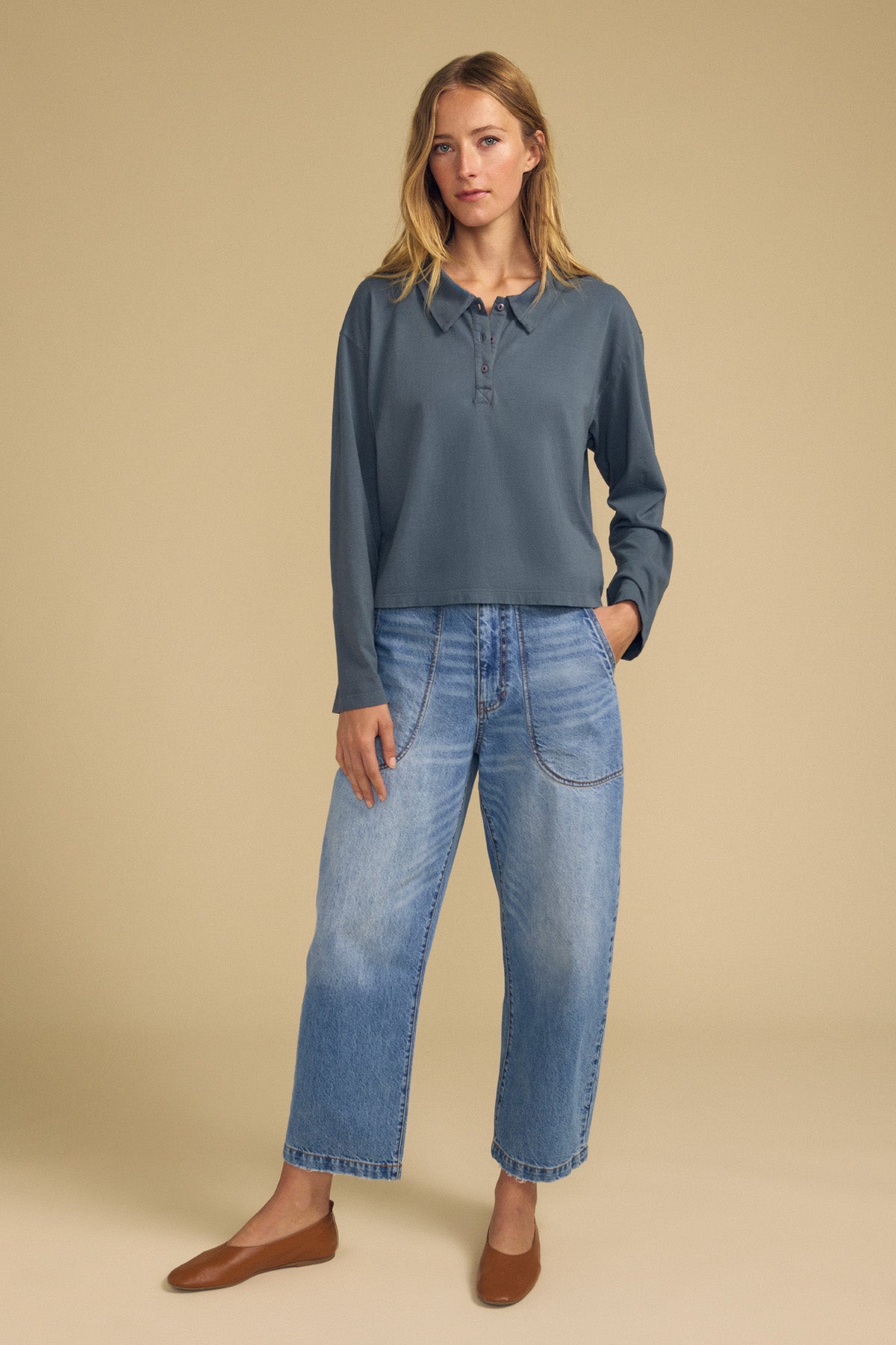 A person stands against a beige background wearing the NILLI POLO by Velvet by Graham & Spencer—a blue long-sleeve vintage cotton jersey top with a boxy fit and drop shoulder—paired with wide-leg blue jeans featuring patch pockets and brown flats.