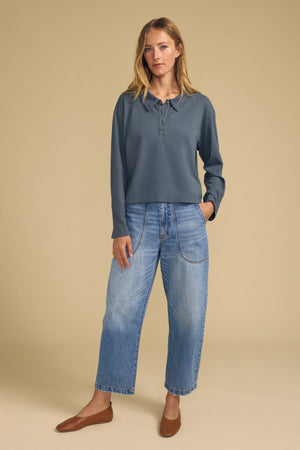 A person stands against a beige background wearing the NILLI POLO by Velvet by Graham & Spencer—a blue long-sleeve vintage cotton jersey top with a boxy fit and drop shoulder—paired with wide-leg blue jeans featuring patch pockets and brown flats.