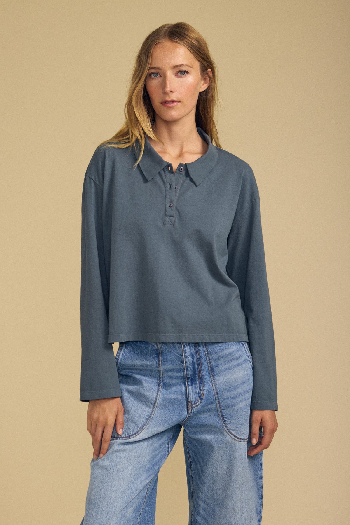 A woman wearing the NILLI POLO long-sleeve, drop-shoulder, buttoned-collar grey shirt by Velvet by Graham & Spencer and loose blue jeans stands against a beige background.-41964690669761