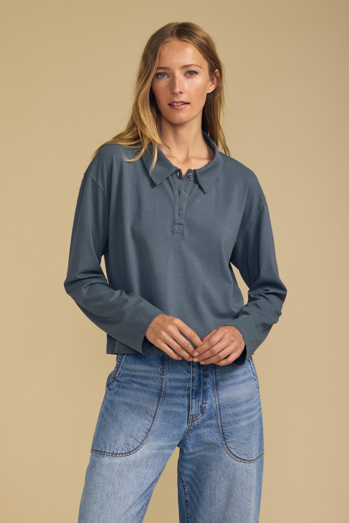 A woman stands against a beige background wearing Velvet by Graham & Spencer’s NILLI POLO—a long-sleeved, slate-blue boxy fit polo crafted from vintage cotton jersey—paired with light blue jeans.