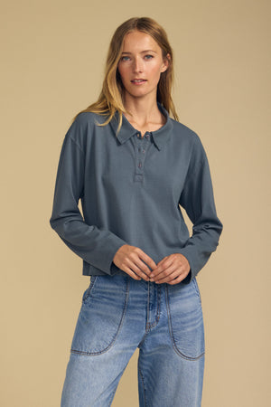 A woman stands against a beige background wearing Velvet by Graham & Spencer’s NILLI POLO—a long-sleeved, slate-blue boxy fit polo crafted from vintage cotton jersey—paired with light blue jeans.