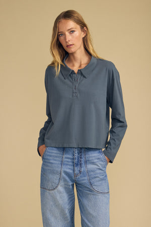 A woman stands against a plain beige background with her hands in her pockets, wearing the Velvet by Graham & Spencer NILLI POLO—a long-sleeve, buttoned gray boxy-fit polo crafted from vintage cotton jersey—paired with high-waisted blue jeans.