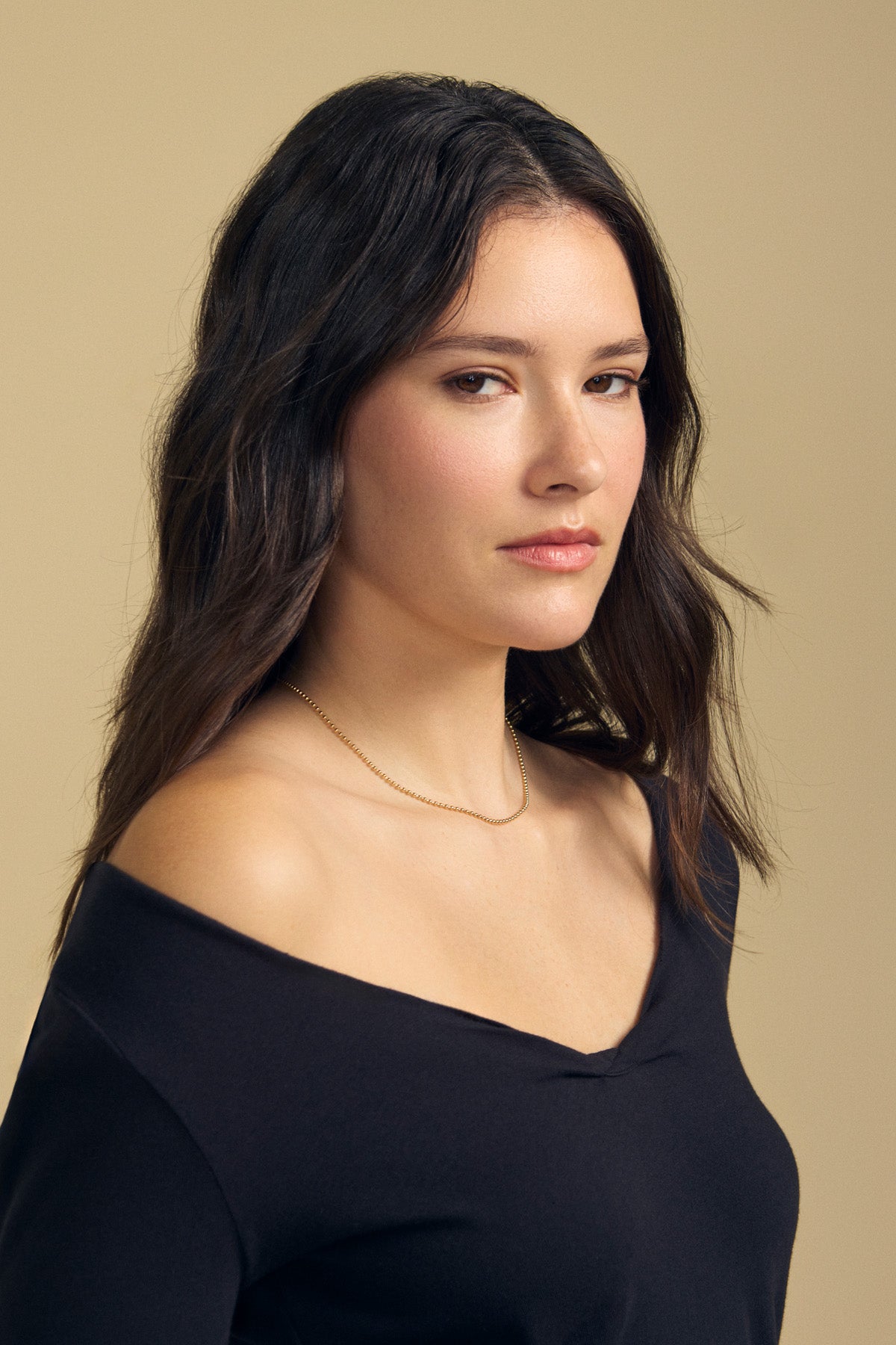 A woman with long dark hair, wearing the NOURA TEE by Velvet by Graham & Spencer—a soft cotton jersey long sleeve top with a scoop V-neckline—paired with a gold necklace, poses against a beige background.