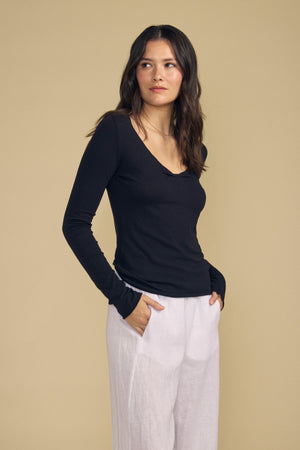 A woman with wavy dark hair stands with her hands in her pockets, looking to the side against a beige background. She is wearing the NOURA TEE by Velvet by Graham & Spencer, a black long-sleeve cotton jersey top featuring a scoop V-neckline, paired with white pants.