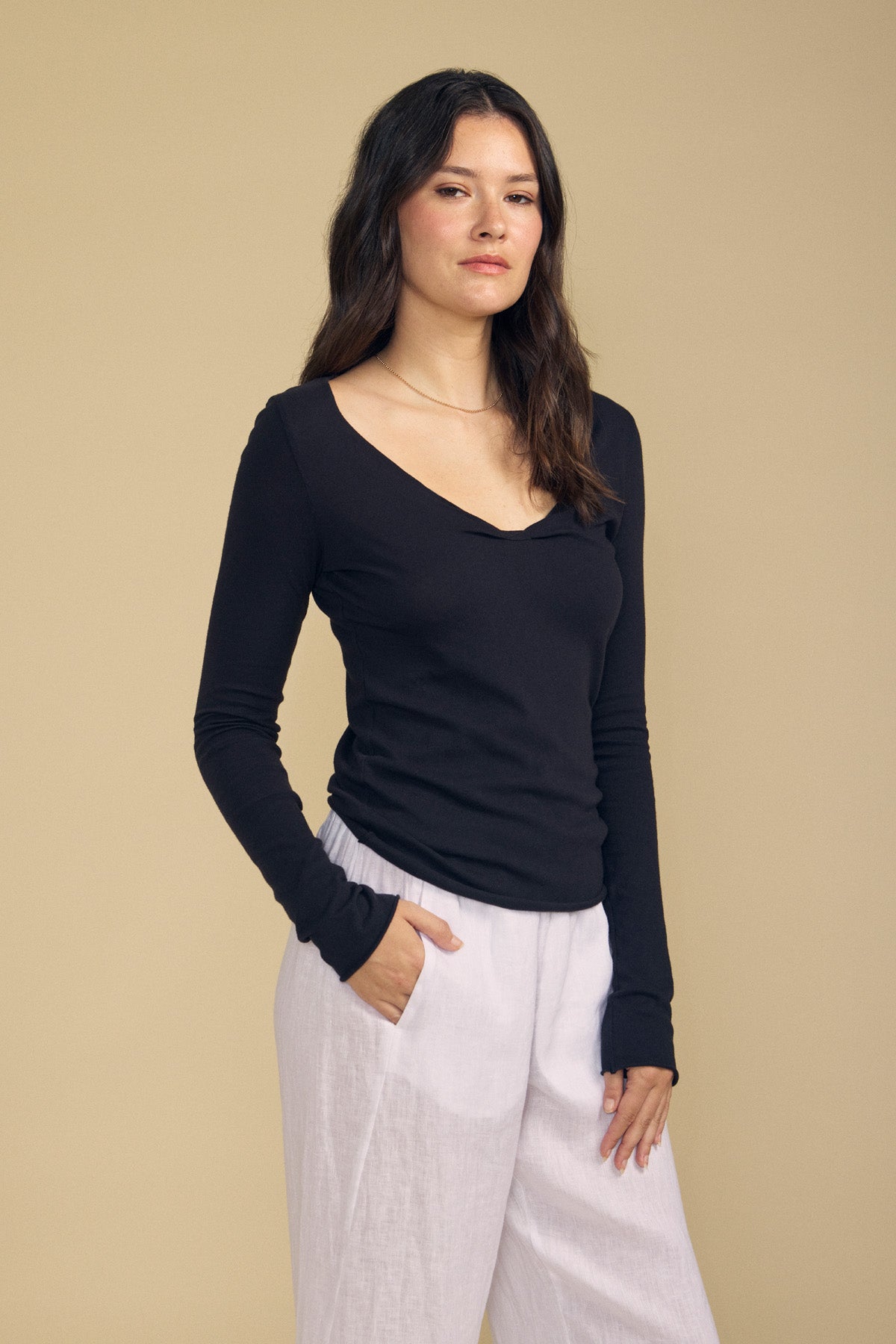 A woman with long dark hair, wearing the Velvet by Graham & Spencer NOURA TEE—a black cotton jersey top featuring a scoop V-neckline—paired with white pants, stands against a plain beige background and looks at the camera.-41947075805377