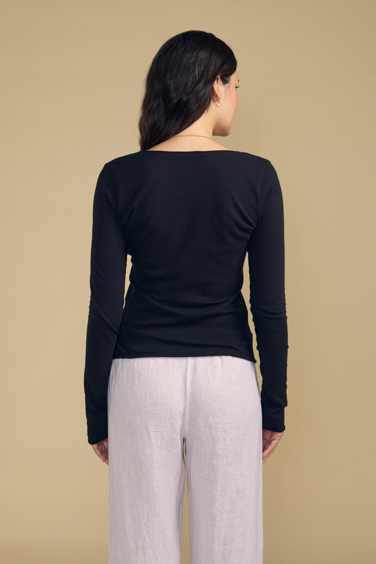 A woman with long dark hair, dressed in the Velvet by Graham & Spencer NOURA TEE—a black, long-sleeve cotton jersey top—and white pants, stands with her back to the camera against a beige background.
