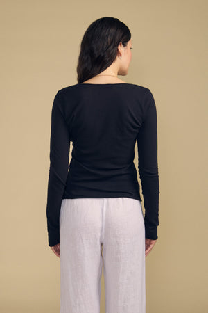 A woman with long dark hair, dressed in the Velvet by Graham & Spencer NOURA TEE—a black, long-sleeve cotton jersey top—and white pants, stands with her back to the camera against a beige background.