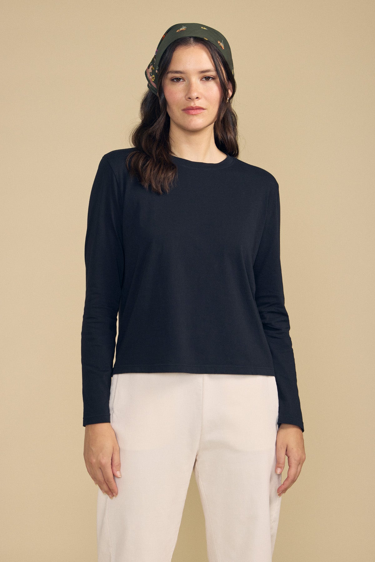 A woman wearing the VICKY TEE by Velvet by Graham & Spencer—a black cotton jersey long-sleeve—paired with white pants and a dark patterned headscarf stands against a plain beige background, creating an effortlessly stylish look ideal for everyday layering.