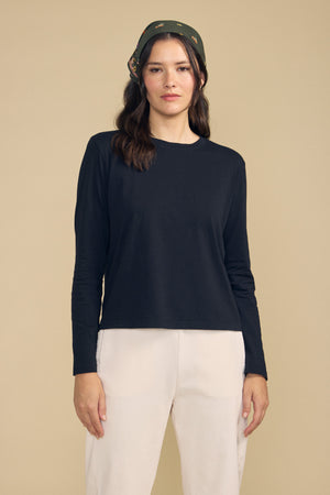 A woman wearing the VICKY TEE by Velvet by Graham & Spencer—a black cotton jersey long-sleeve—paired with white pants and a dark patterned headscarf stands against a plain beige background, creating an effortlessly stylish look ideal for everyday layering.