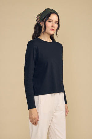 A woman stands against a beige background, wearing the Velvet by Graham & Spencer VICKY TEE—a black cotton jersey long-sleeve top with a classic crew neckline—paired with light-colored pants and a patterned headscarf, looking at the camera with a neutral expression.