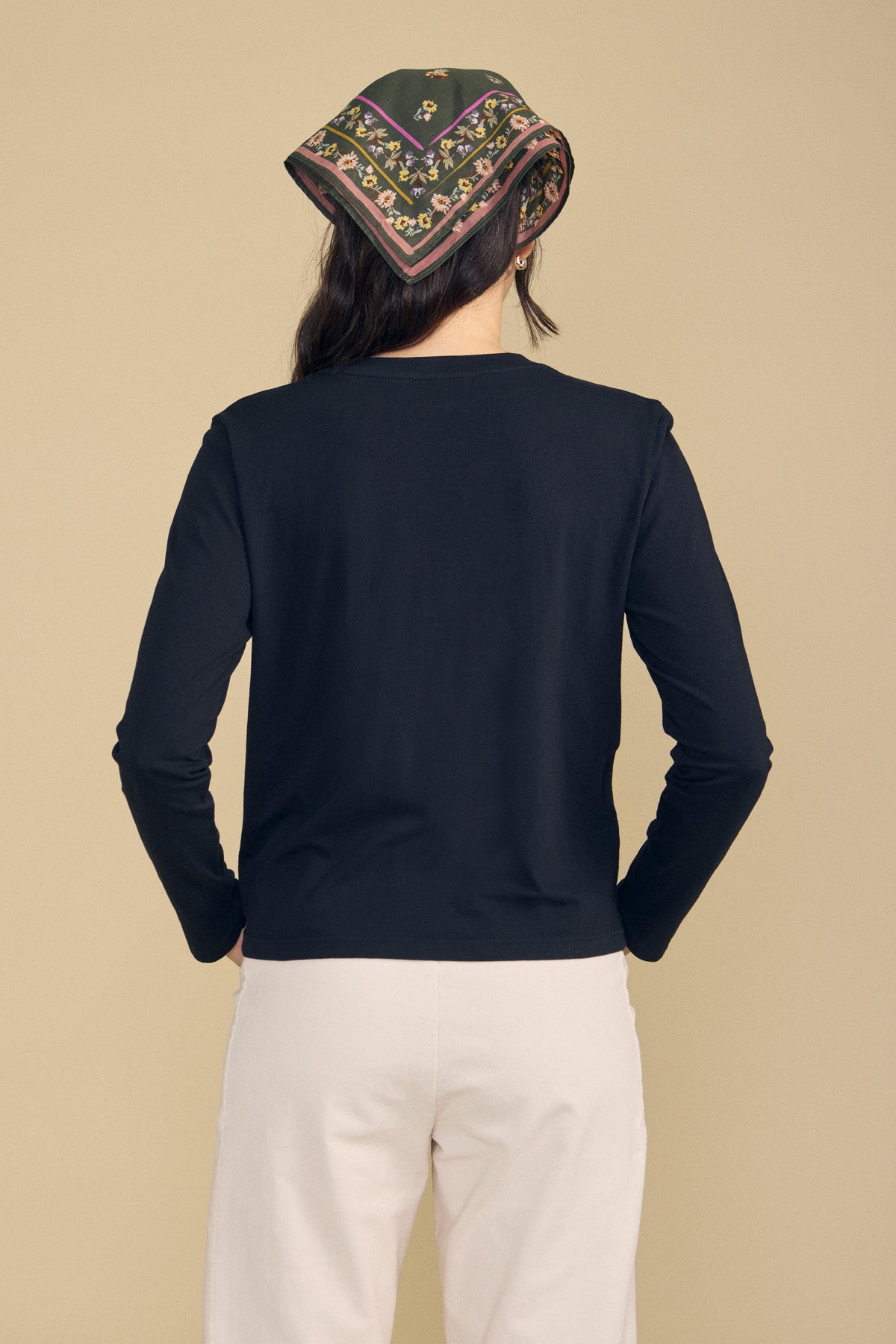 A person stands facing away, wearing the VICKY TEE by Velvet by Graham & Spencer—a black cropped cotton jersey tee—paired with light pants and a patterned headscarf against a beige background, creating an effortless look perfect for everyday layering.
