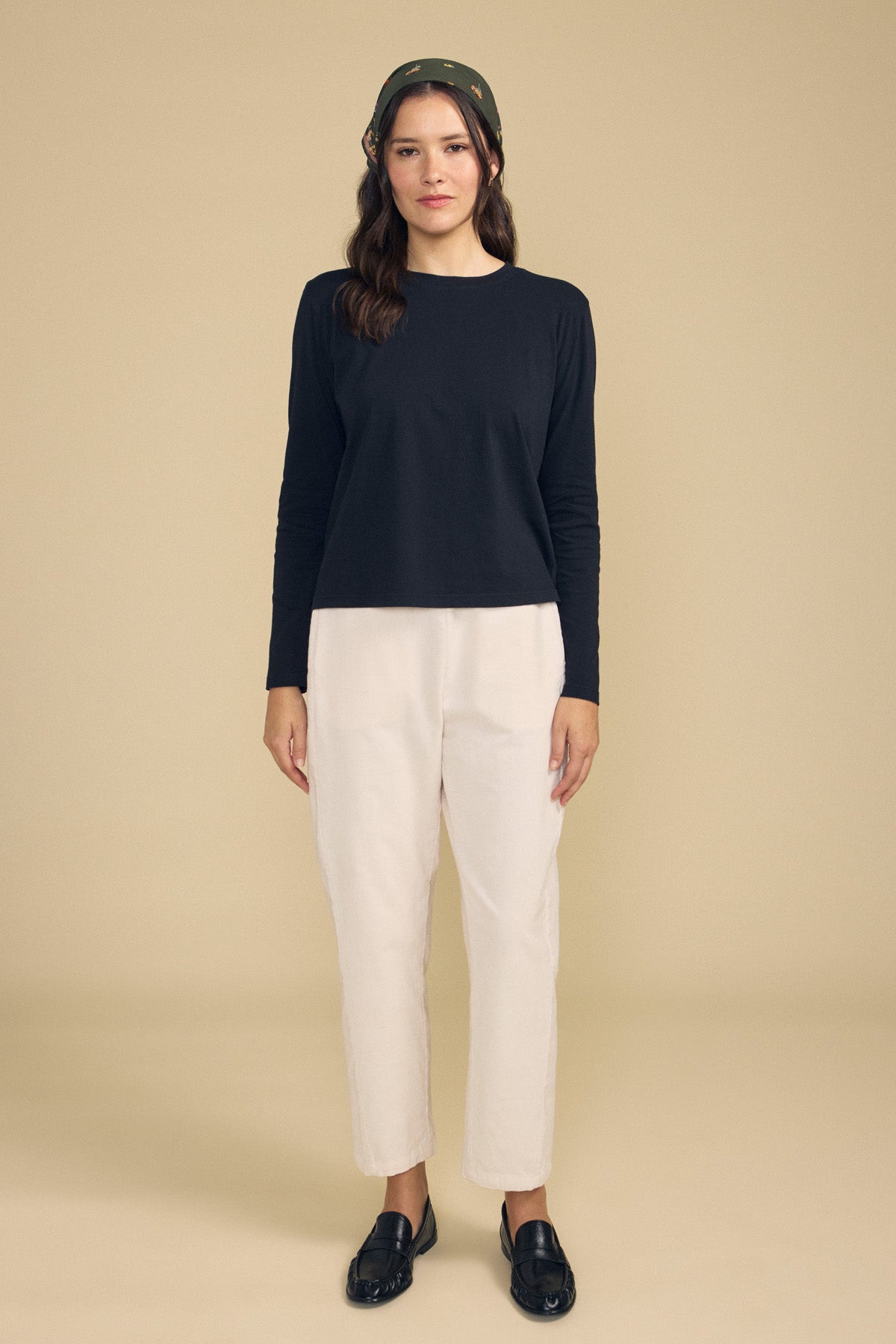 A woman stands facing forward against a plain beige background, wearing a black long-sleeve shirt, black loafers, a dark headscarf, and the LANEY CORDUROY PANT by Velvet by Graham & Spencer—a light-colored pinwale corduroy cropped pant with an elastic waist.