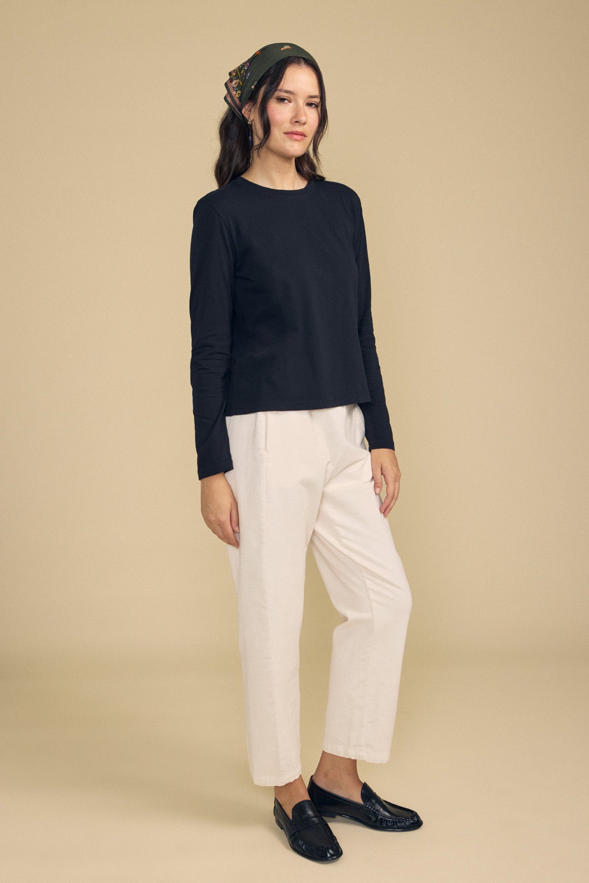 Woman stands against a beige background wearing a black long-sleeve top, the LANEY CORDUROY PANT by Velvet by Graham & Spencer in cream with an elastic waist and cropped fit, black loafers, and a patterned headscarf.-41860486070465
