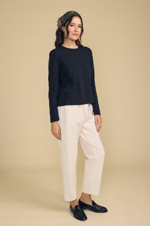 Woman stands against a beige background wearing a black long-sleeve top, the LANEY CORDUROY PANT by Velvet by Graham & Spencer in cream with an elastic waist and cropped fit, black loafers, and a patterned headscarf.