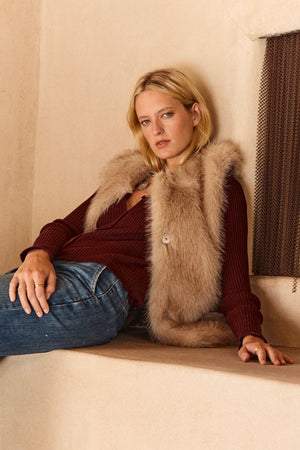 A woman wears the ANASTASIA FAUX FUR VEST by Velvet by Graham & Spencer over a maroon sweater and blue jeans, sitting casually against a beige wall and looking at the camera with a neutral expression.