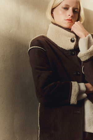 A person with short blond hair wears the SHONA SHERPA REVERSIBLE JACKET by Velvet by Graham & Spencer over a light sweater, standing against a beige wall.