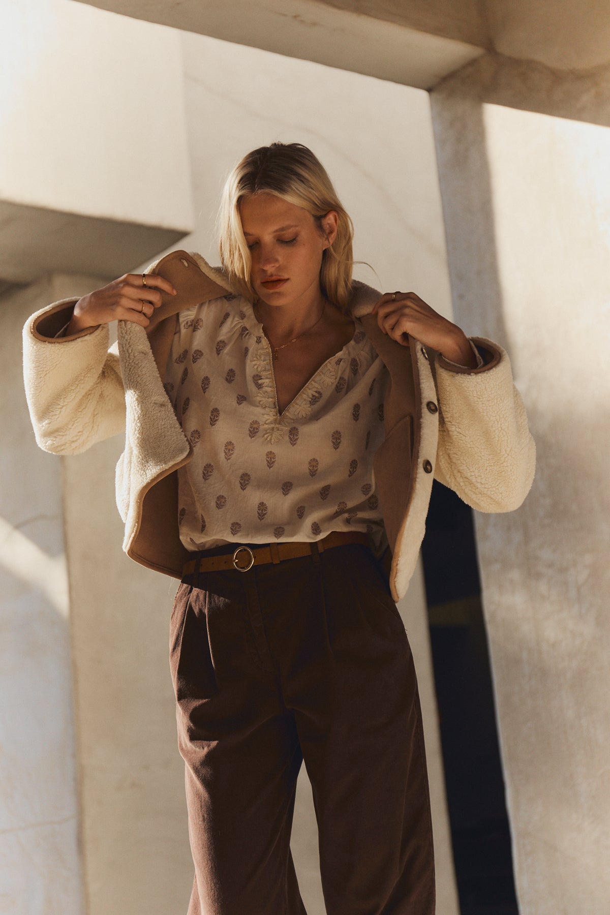   A woman stands under architectural structures, wearing a beige patterned shirt, brown pants, and the AMARA SHERPA REVERSIBLE JACKET by Velvet by Graham & Spencer draped over her shoulders. 