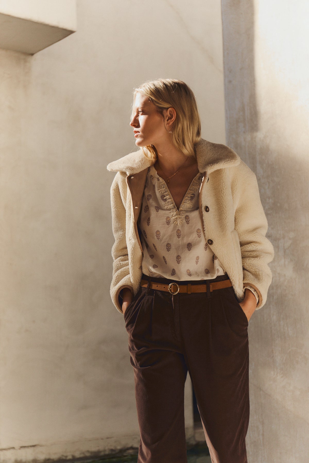   A woman stands with hands in pockets, wearing the AMARA SHERPA REVERSIBLE JACKET by Velvet by Graham & Spencer, over a patterned blouse and brown pants, gazing to the side in natural light against a plain background. 