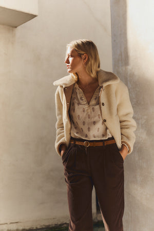 A woman stands with hands in pockets, wearing the AMARA SHERPA REVERSIBLE JACKET by Velvet by Graham & Spencer, over a patterned blouse and brown pants, gazing to the side in natural light against a plain background.