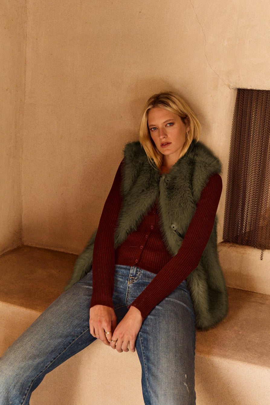 A woman wearing the ANASTASIA FAUX FUR VEST by Velvet by Graham & Spencer over a red sweater and blue jeans sits on a beige bench by a fireplace, highlighting this cozy layering piece for your cold-weather style.-39815116226753