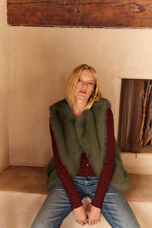 A woman with blonde hair sits on a ledge in a rustic indoor setting, wearing the ANASTASIA FAUX FUR VEST by Velvet by Graham & Spencer—a cozy layering essential for your cold-weather wardrobe.