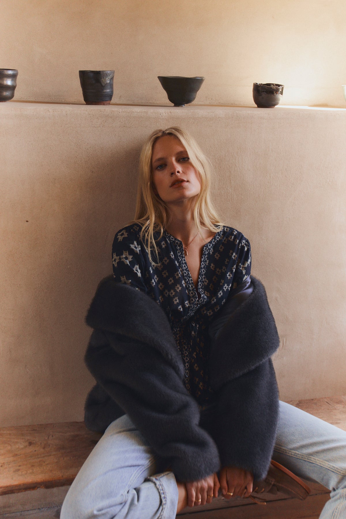 A woman with long blonde hair sits on the floor by a beige wall, wearing the CHARLOTTE TOP by Velvet by Graham & Spencer with smocked elastic cuffs, a dark fur coat, and light blue jeans; ceramic bowls rest on a ledge above her.-39815174062273