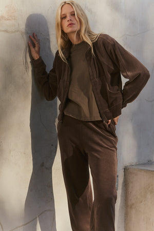 A person with long blonde hair stands against a light concrete wall, wearing the CLAIRE JACKET by Velvet by Graham & Spencer—an easy-fitting brown cotton velour hoodie paired with matching pants and a brown shirt.