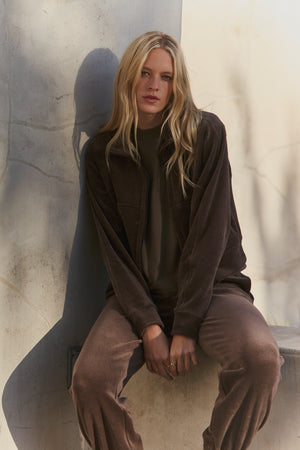 A person with long blonde hair, wearing the CLAIRE JACKET by Velvet by Graham & Spencer and matching pants, sits on a concrete ledge against a light gray wall.