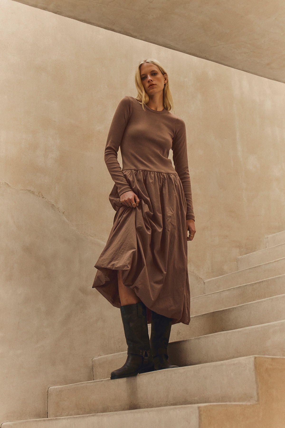 A woman wearing the DOLCE DRESS by Velvet by Graham & Spencer stands on light-colored stairs in black boots against a beige background.