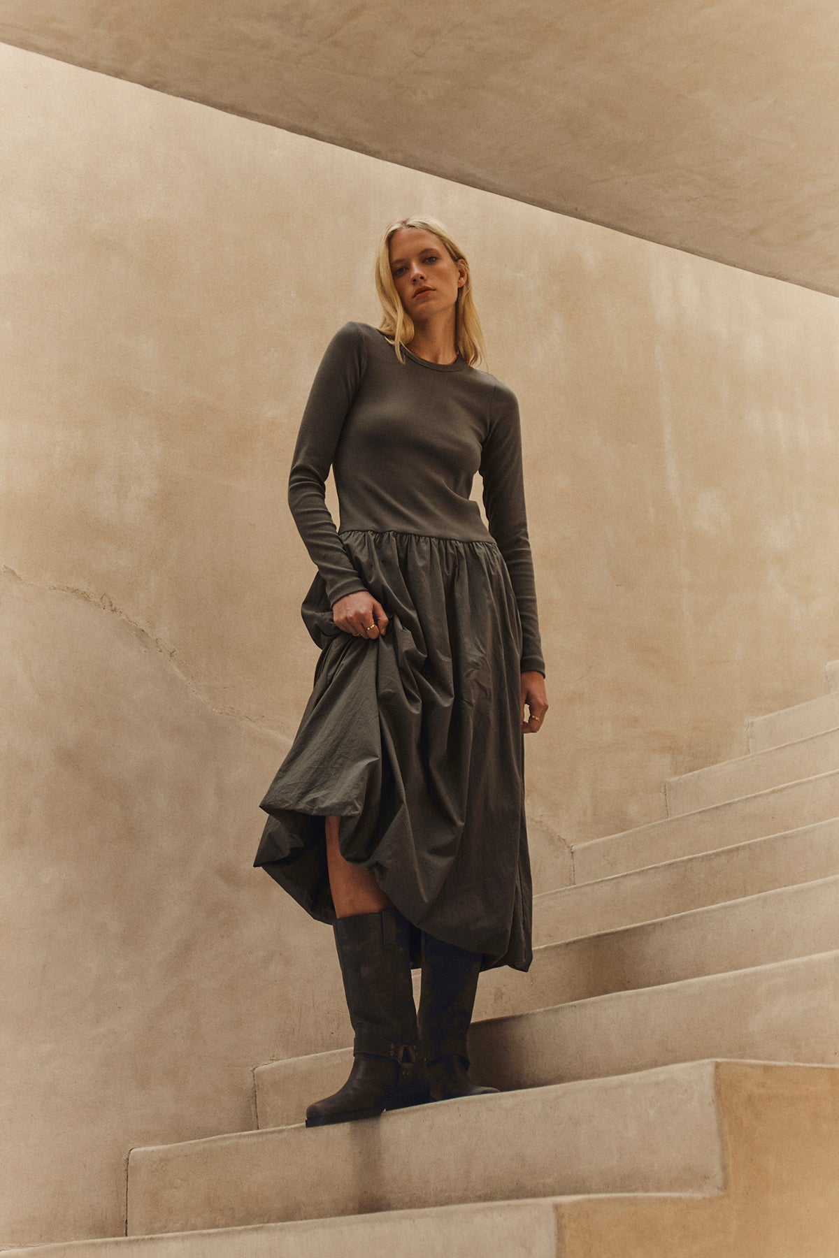 A person with long blonde hair stands on curved concrete steps, wearing the DOLCE DRESS by Velvet by Graham & Spencer and dark boots, set against a beige wall.-39559481360577