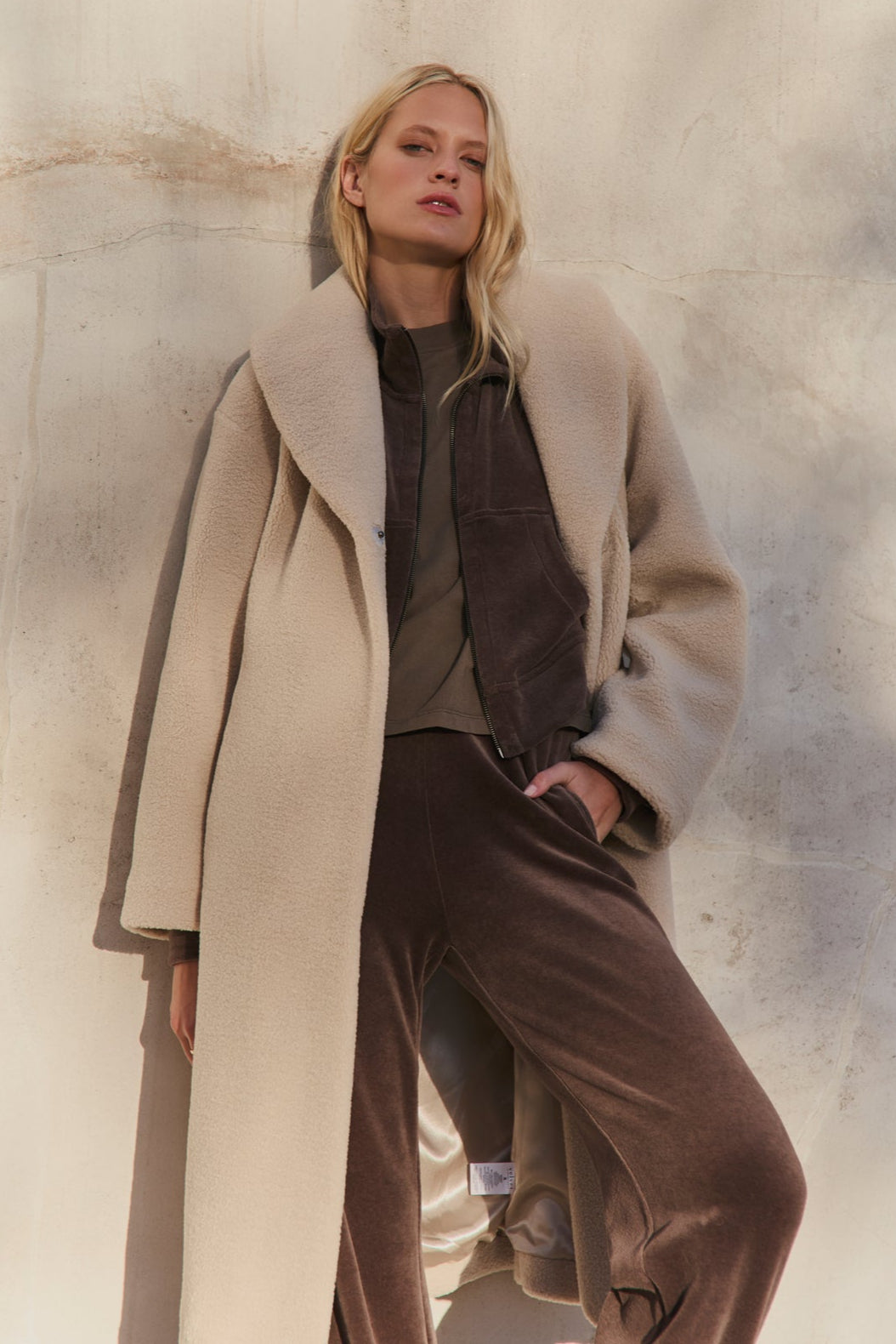 A woman stands against a textured wall wearing the Velvet by Graham & Spencer ELEANOR SHERPA REVERSIBLE COAT over a brown jacket and matching pants, with one hand in her pocket.-39838347952321