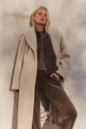 A woman stands against a textured wall wearing the Velvet by Graham & Spencer ELEANOR SHERPA REVERSIBLE COAT over a brown jacket and matching pants, with one hand in her pocket.