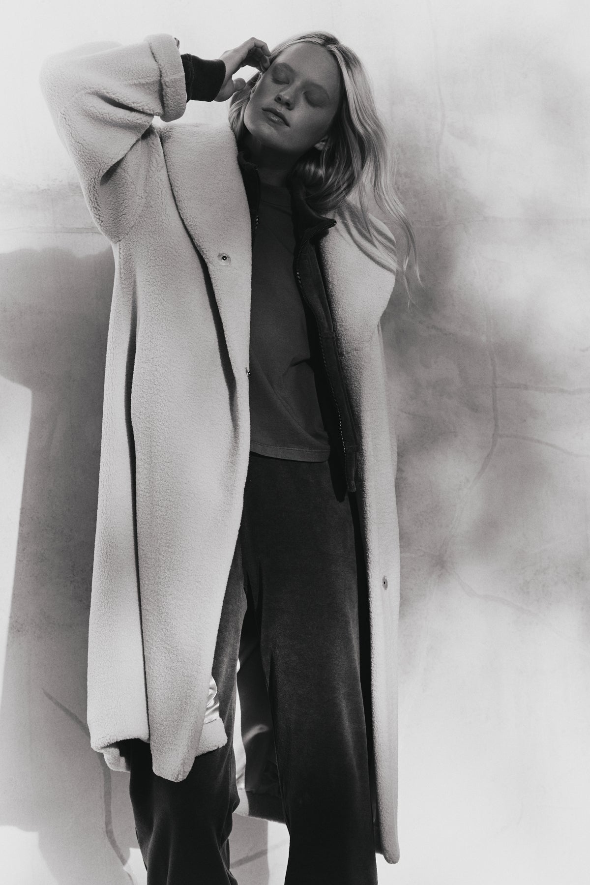 Black and white photo of a person standing against a textured wall, wearing the ELEANOR SHERPA REVERSIBLE COAT by Velvet by Graham & Spencer and loose pants, raising one hand to their head.-39838347886785